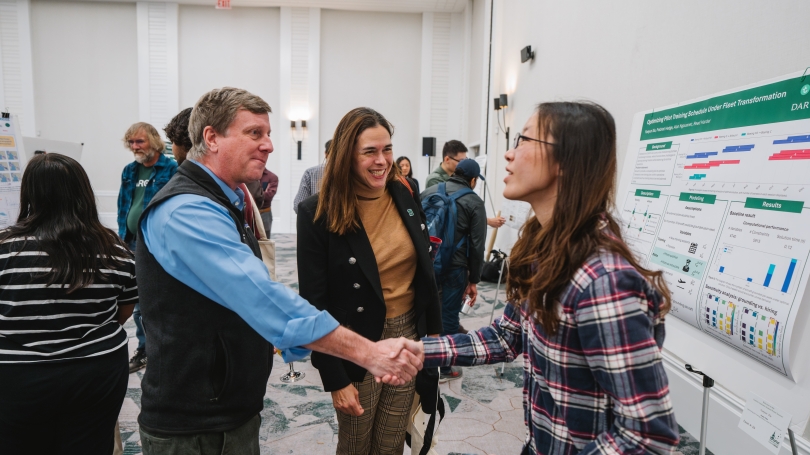a man and woman standing in front of a poster shake hands while another woman stands between them smiling.