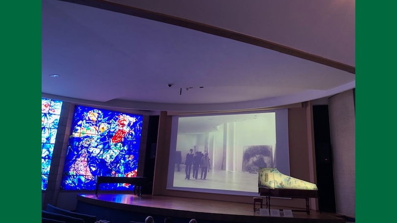 An art exhibit with blue stained glass images and a stage with a piano and a projected image